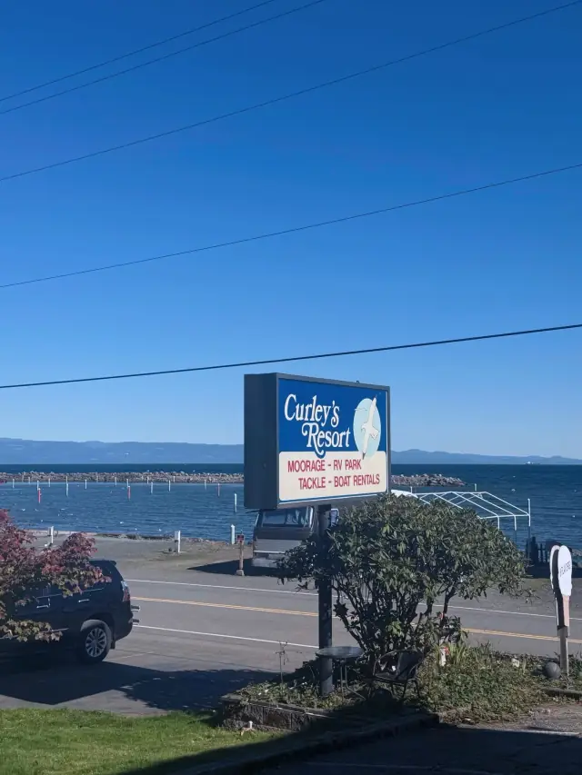 The Curley's Resort roadside sign on Front Street listing moorage, RV park and tackle, with the Strait behind it